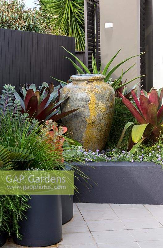 A large amphora shaped water bowl in a raised garden bed with a large bromeliad next to a group of cylindrical pots planted with a variety of plants.