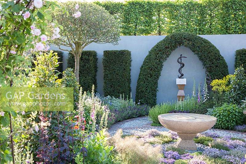 Contemporary Apothecary Garden with large water-basin surrounded by herb-lawn of Thymus serpyllum - Thyme. Umbrella-pruned Cratageus - Hawthorn, Myrtus communis - Myrtle. Taxus baccata - Yew topiary around sculpture of Aesclepius' staff, the symbol of healing. Rosa - Rose and Laurus nobilis - Bay