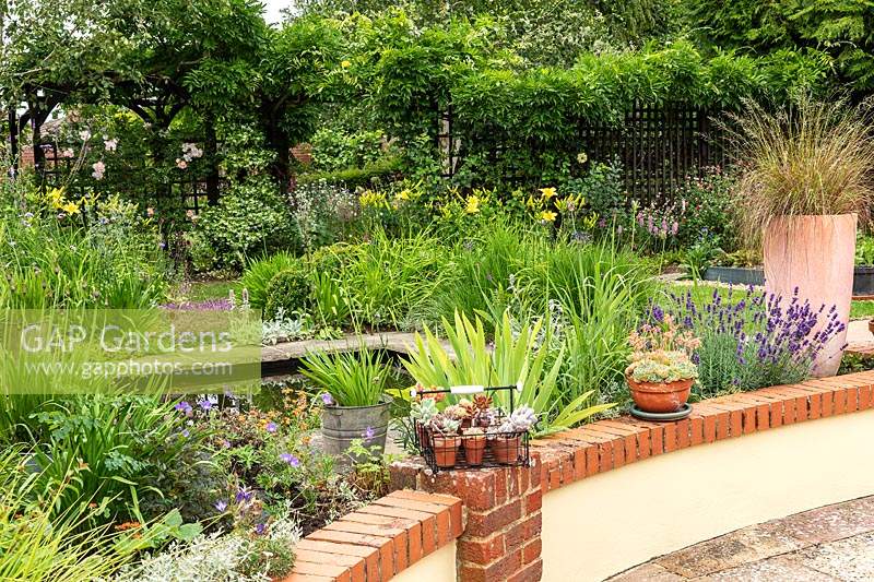 A small informal suburban country garden with a pond, shrubs and perennials with climbers on a boundary fence, view over low patio wall 