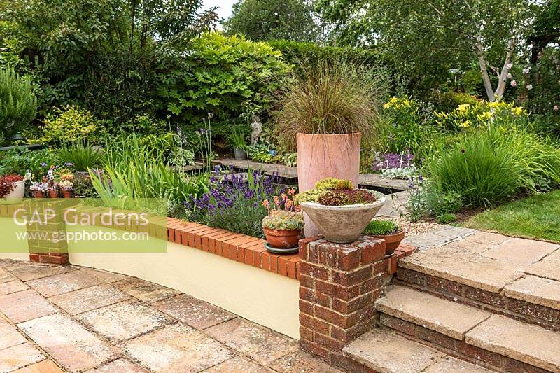 A sunken patio with a low retaining wall, containers and stone steps leading up to a pond and informal country garden planting