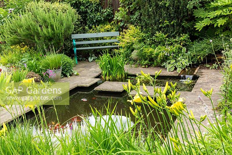 A small garden pond with stepping stones in a suburban country garden, surrounded with stone paving, a garden seat and informal mixed planting of perennials, such as Hemerocallis, and shrubs