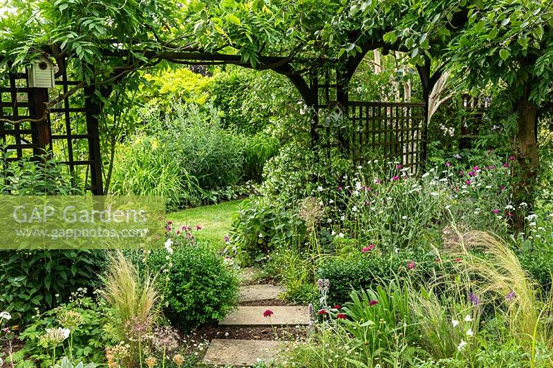 Stepping stones lead under the arch of a wooden pergola with bird box. Borders of perennials and grasses in an informal cottage-garden style