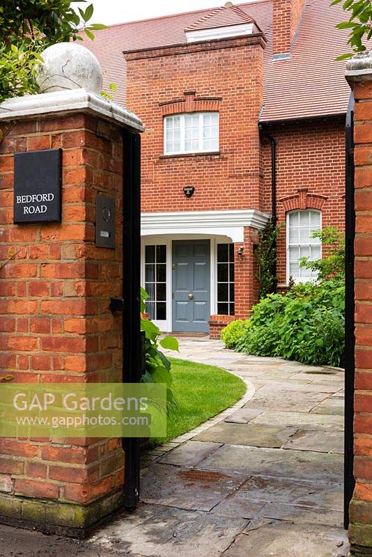 Entrance to house through black painted gate, curved paved path to front door, boundary brick wall matches house bricks