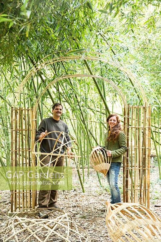 Man and a woman holding bamboo craft objects under a bamboo arch in a clearing of living Phyllostachys - Bamboo