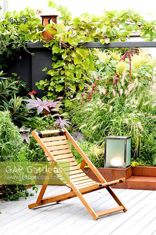 Relaxed seating area beside a border wall created to hide the adjacent building. Corten containers planted with chard, pennisetum, amaranths and castors, rosemary, tulbaghie