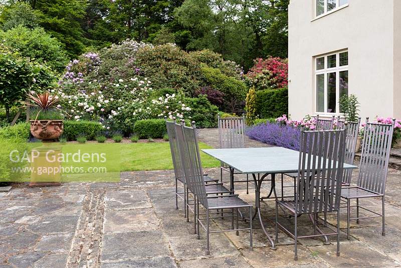 Table and chairs set up on terrace, surrounded by flowering borders.
