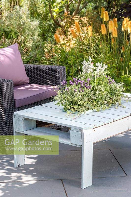 Wooden pallet table with integral sunken  planter with summer planting on slate patio in summer accompanied by outdoor chairs.