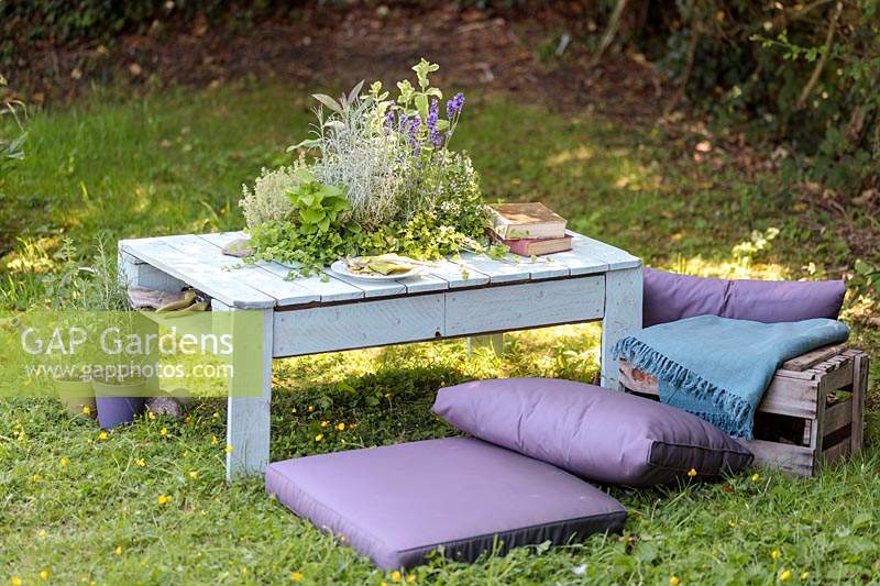 Table made out of a wooden pallet, with central planter planted with mixed herbs in summer country garden with cushions. 