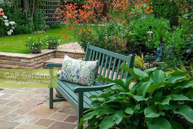 Paved patio with garden bench with floral pattern cushion and Hosta in container against a raised bed of orange Geum and other perennials. Steps lead from patio to a small lawn - Open Gardens Day, Drinkstone, Suffolk