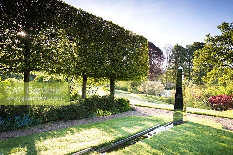 Stainless steel obelisk at the end of water rill framed by pleached hornbeams in May