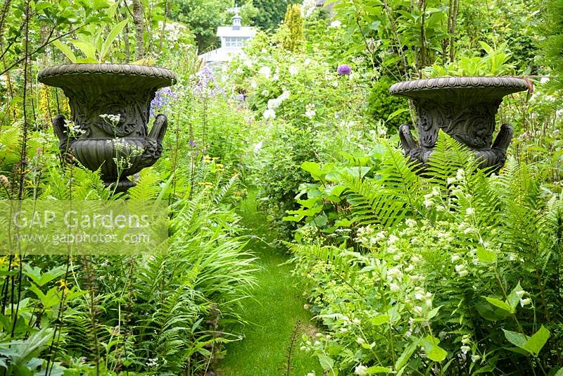 Decorative urns in borders packed with plants either side of grass path. 