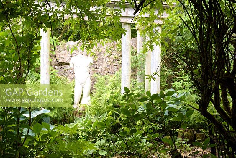 Classical statue framed by temple columns and lush planting including ferns and hostas in the walled garden 