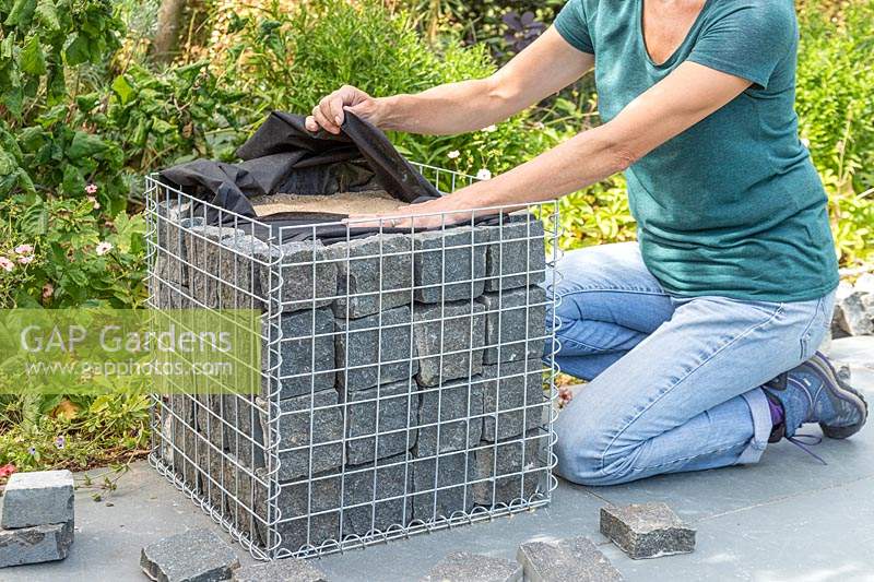 Woman folding over excess liner at the top of gabion basket lined with granite blocks and sand. 