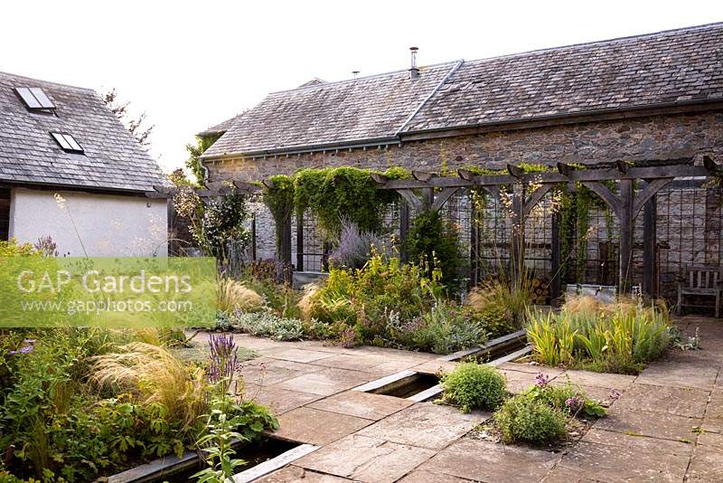 Courtyard garden with timber pergola, rill and planting including Stipa tenuissima, S. gigantea, Phlomis russeliana, Perovskia 'Blue Spire', irises,  Verbena hastata and silvery stachys at Am Brook Meadow, Devon in August. 