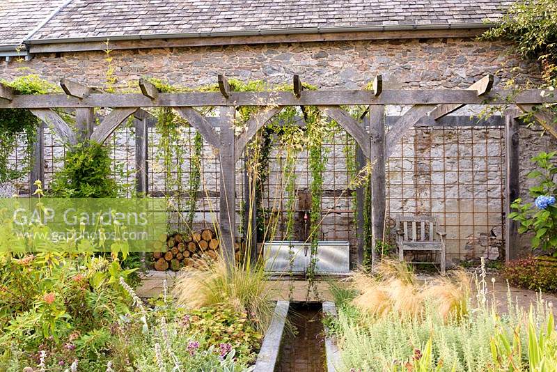 Courtyard garden with timber pergola, rill, galvanised trough water feature and planting including Stipa tenuissima, S. gigantea, Phlomis russeliana, irises and silvery stachys at Am Brook Meadow, Devon in August. 