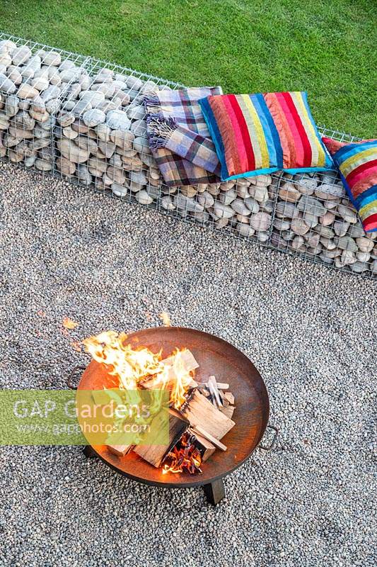 Lit firepit area with colourful cushions on gabion benches and pebble surface