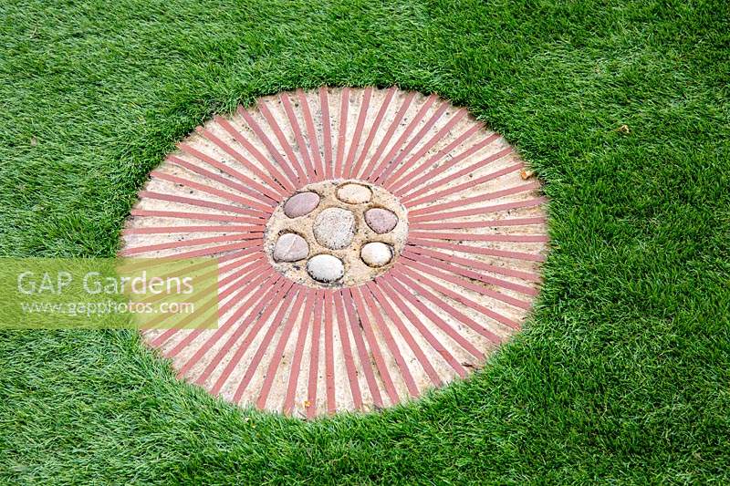 Circular terracotta path step with pebbles at centre set in artificial lawn