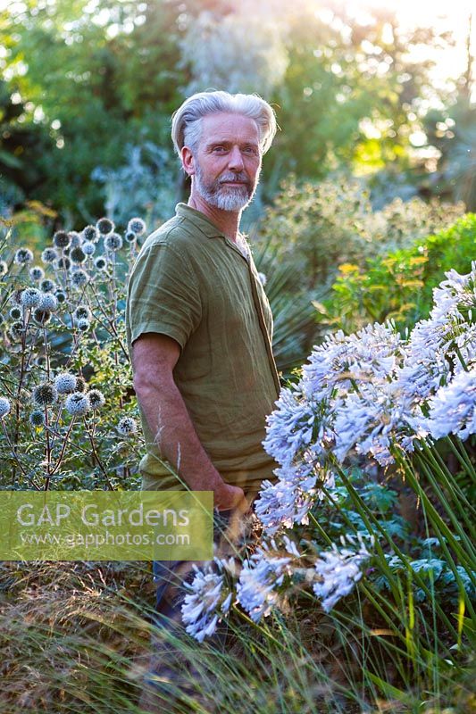Nick Macer in the garden with Agapanthus 'Windsor Grey' in foreground, Pan Global Plants, Frampton on Severn, Gloucestershire, UK.