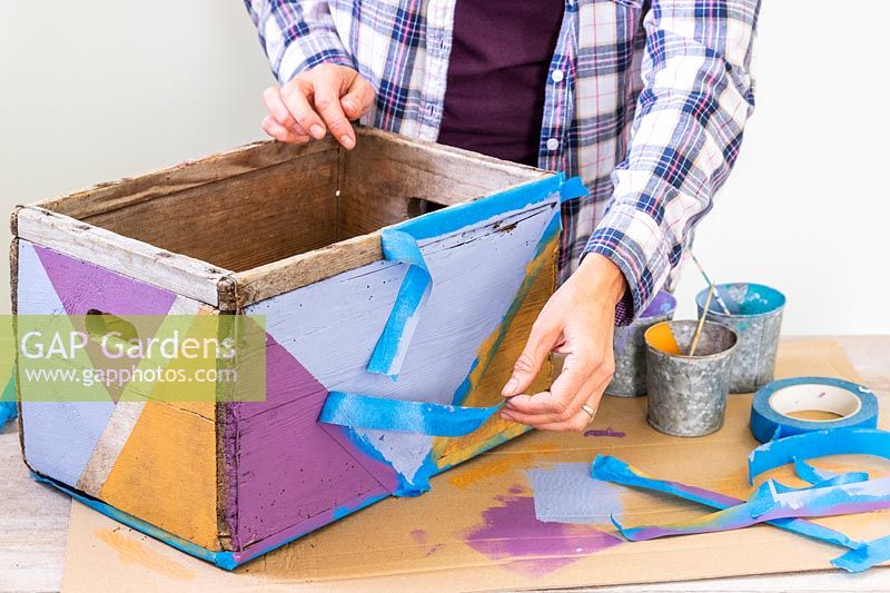 Woman removing masking tape to reveal wooden box painted in a multicoloured geometric pattern