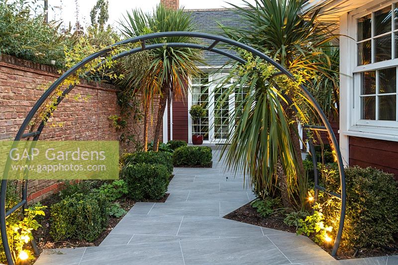 View through circular metal arch to paved area by house, beds with Cordyline and lighting 