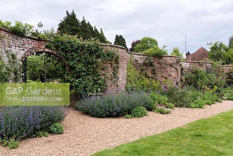 A serpentine or crinkle crankle wall with gated archway and gravel path and long border planted with nepeta, euphorbia and allium