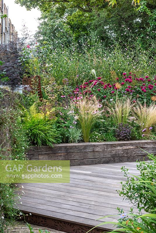 Decked courtyard garden surrounded by raised beds planted with  sun-loving, wildlife friendly perennials and grasses, with view of cityscape beyond.
