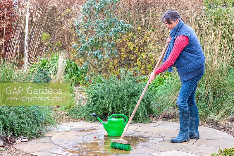 Woman using a brush to scrub a patio after having applied a mixture of white vinegar and water to clean the paving