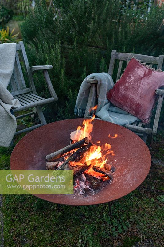 A lit fire bowl made from rusted corten steel,  wooden garden chairs with wool rugs and a cushion by a large rosemary bush.