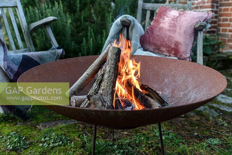 A lit fire bowl made from rusted corten steel, wooden garden chairs with wool rugs and a cushion 