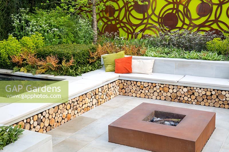 Large sawn Yorkstone stone patio with corten steel fire pit and a bench with colourful cushions and log storage underneath.The Sunken Retreat. RHS Malvern Spring Festival, 2016. Design: Ann Walker for Graduate Gardeners
