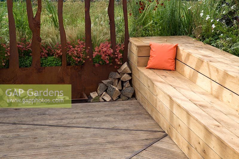 A timber framed wooden bench seating area with orange cushion, by firepit with logs and a corten steel screen divider. Prospect and Refuge garden, RHS Tatton Park Flower Show, 2017. Designer: Anca Panait