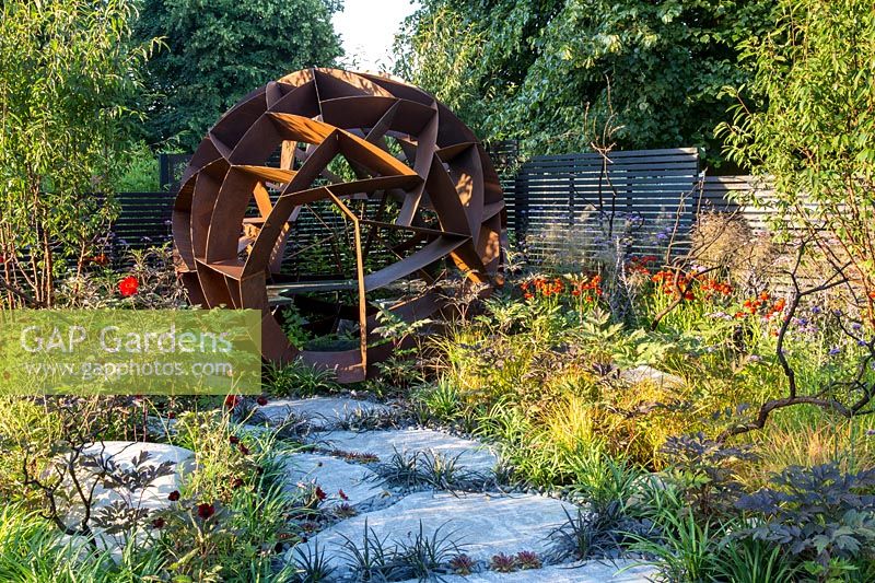 Large Caledonian stone paving slabs path interplanted with Ophiopogon planscapus 'Nigrescens' and Sempervivums  leading to a Corten steel sphere by William Roobrouck - borders planted with Cosmos atrosanguineus, Dahlias, ornamental grasses and Helenium 'Moreheim Beauty' Elements Mystique Garden. RHS Hampton Court Flower Show July 2018 Designer: Lawrence Roberts - Sponsor Elements Garden Design 