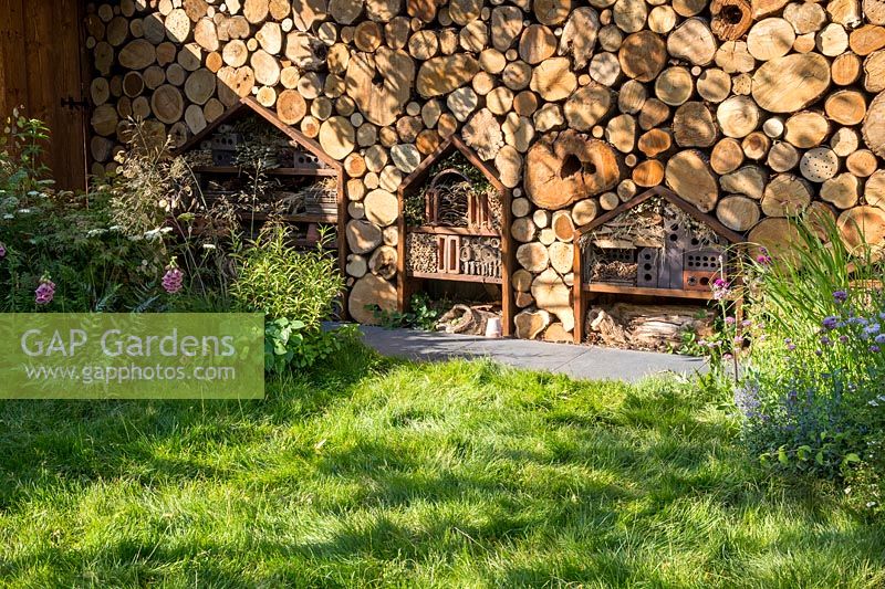 Wildlife and child friendly garden with a wall made from logs habitat for pollinators with insect hotels and an unmown long grass lawn. The Family Garden. RHS Hampton Court Flower Show July 2018  - Designer: Lilly Gomm - Sponsors Practicality Brown, Marshalls, The Tree Company 