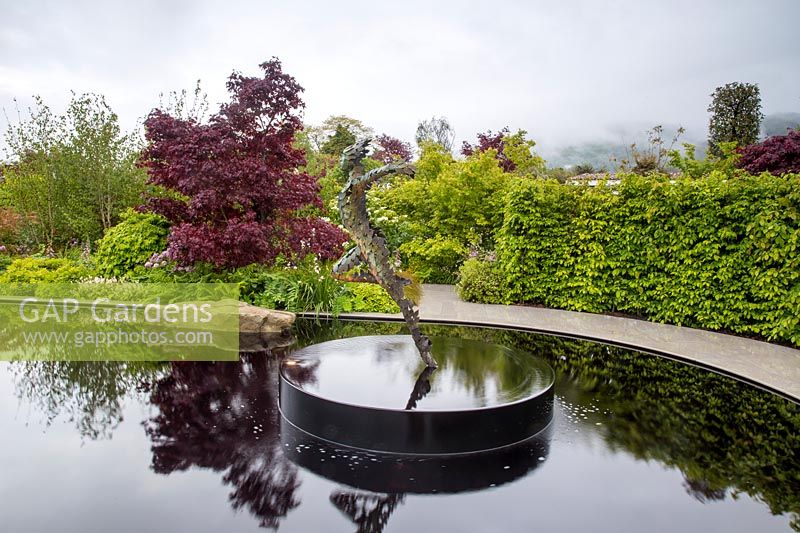 Reflective black pond water feature with sculpture 'Zephyr' by Simon Gudgeon on a misty May spring day - mixed planting - garden hedge and Acer palmatum 'Bloodgood' Garden of Quiet Contemplation. RHS Malvern Spring Festival 2019 - Designer Peter Dowle - Leaf Creative