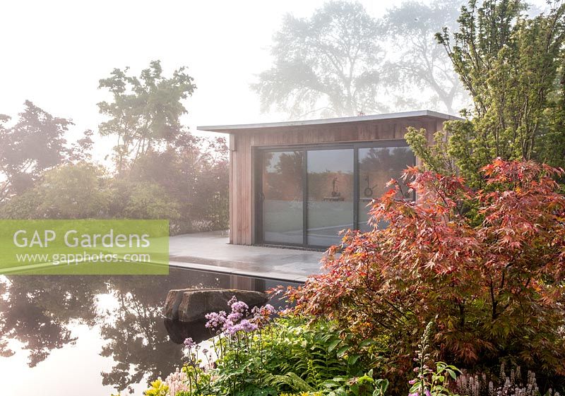 Wooden garden office studio in front of a pool and paved stone patio area on a misty May morning - planted with Acer palmatum 'Shishigashira' in foreground and shrubs. Garden of Quiet Contemplation. RHS Malvern Spring Festival 2019  - Designer Peter Dowle - Leaf Creative 