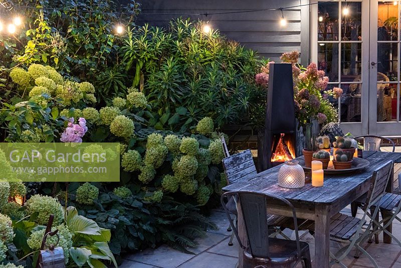 A dining area in a courtyard garden lit by a string of bulbs and candles, heated by a modern outdoor chiminea. Planting behind includes Hydrangea 'Limelight', Rosa 'Blush Noisette' and ferns.