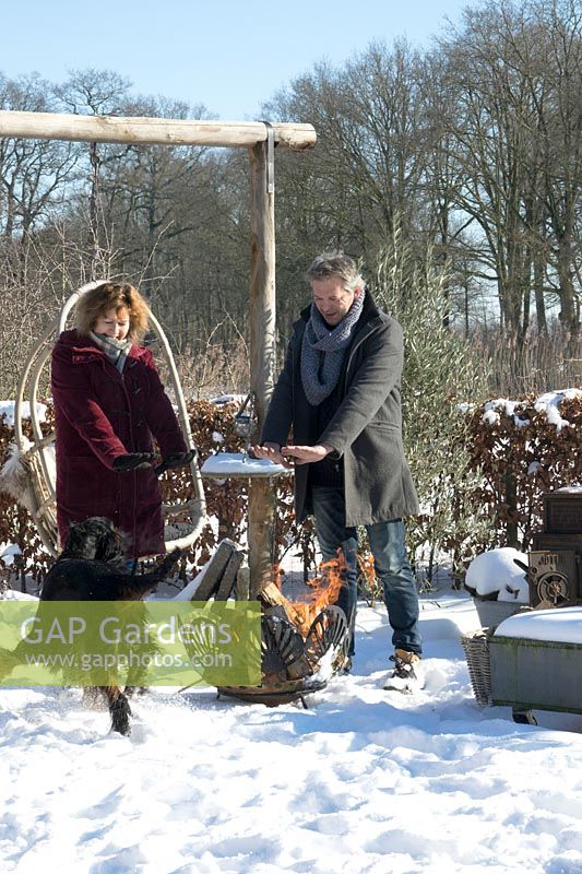 Man and wife, Martijn and Liesbeth warming their hands near the fire basket in the snow with their dog jumping around.