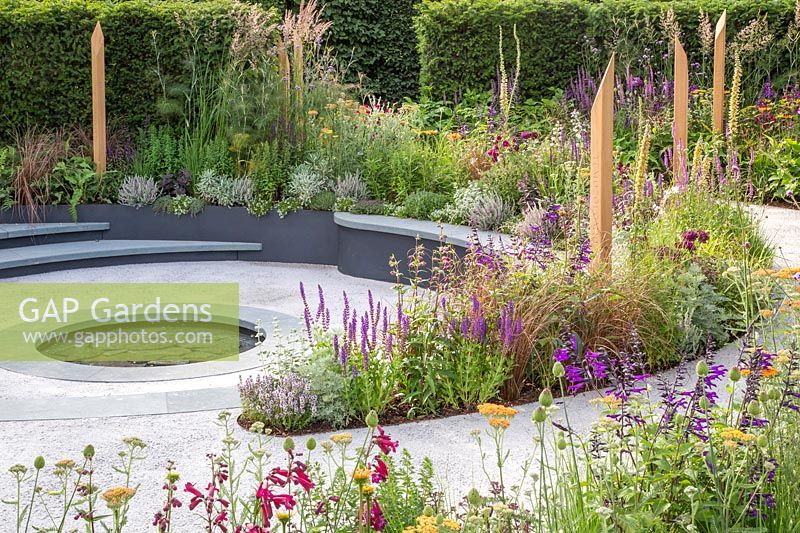 Compacted path spiralling around beds full of colourful summer perennials with inscribed wooden posts leading to central reflection pool. The Cancer Research UK Pledge Pathway to Progress - Hampton Court Flower Festival, 2019.