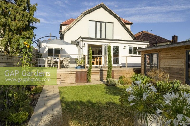 View towards house with seating area on elevated deck and wooden outbuilding in modern family garden 