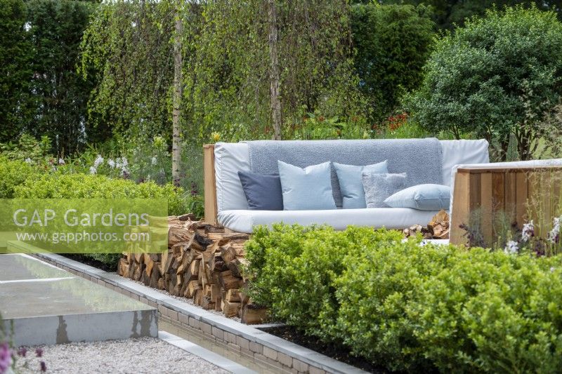 Outdoor seating next to a log pile and water rill, surounded by evergreen hedging - The Viking Friluftsliv Garden - RHS Hampton Court Festival 2021