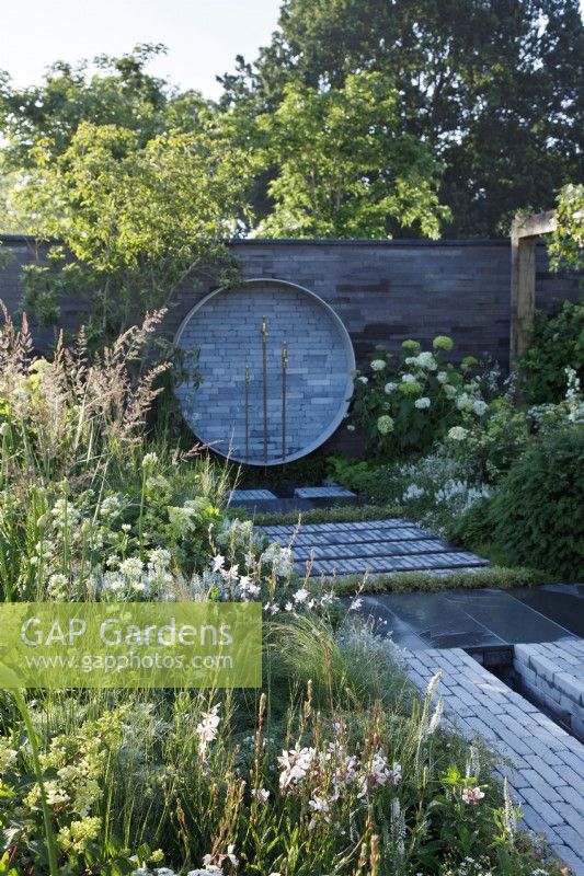 In A place to Meet Again Garden,  the path paved with smooth dutch style pavers cuts through the planting which uses a green and white palette; a water feature of repurposed brass taps stands against a wall of stacked concrete slabs- Designer: Mike Long - Sponsors: Association of Professional Landscapers, Kebur Garden Materials, Creepers Nurseries, Landscape Plus.