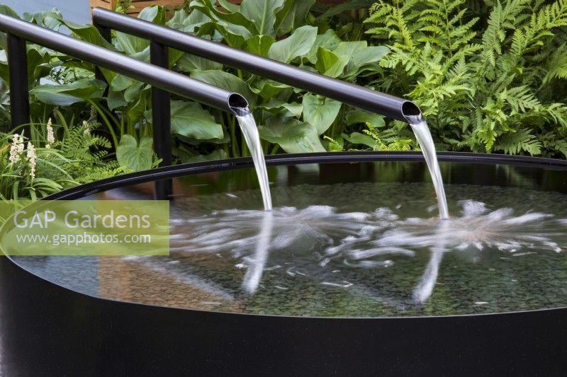 Water flows through dual metal spouts into circular metal raised pool, with plants behind. Calm of Bangkok.