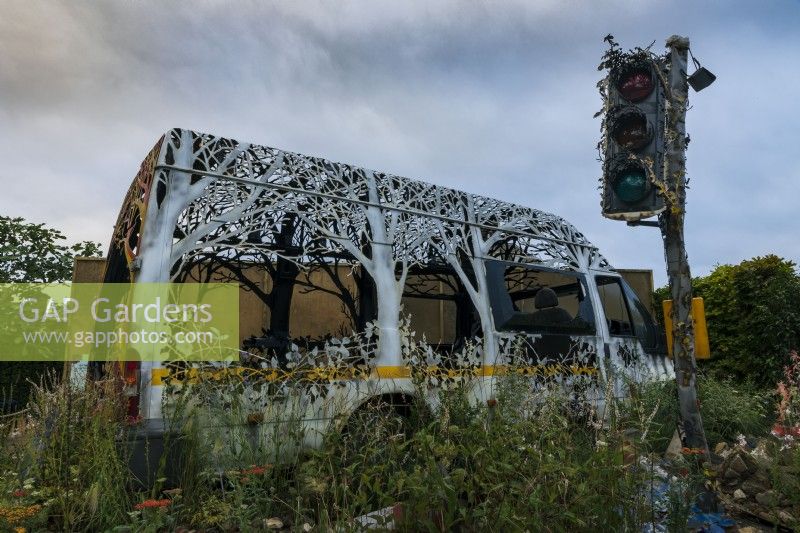 Delays Expected, by Dan Rawlings, exhibited by Saatchi Gallery. Van etched and carved into latticework of trees and plants, surrounded by planting evocative of wasteland, including white Persicaria, achillea, and brambles. Decaying traffic light in foreground.