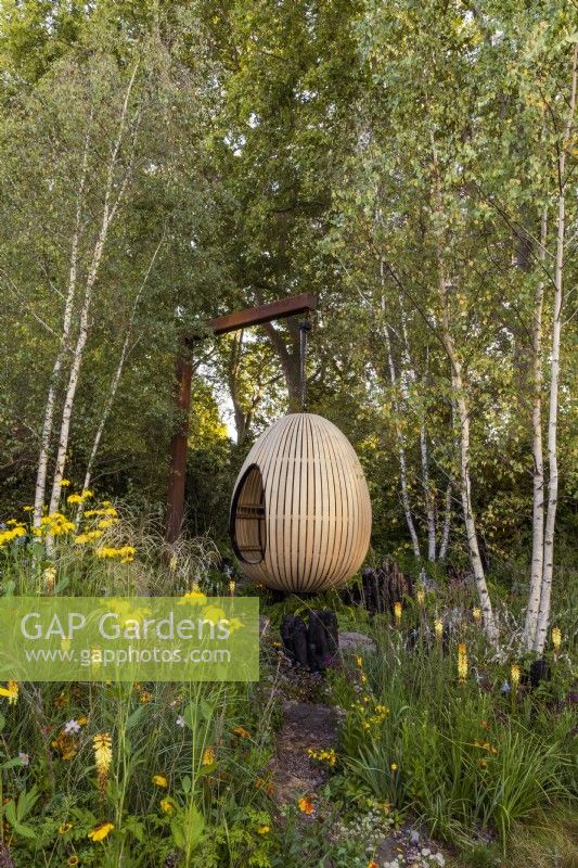 Yeo Valley Organic Garden. A suspended, steam-bent, oak egg seat forms the focal point of the garden. Naturalistic perennial meadow with rough pathway and burnt wood sculptures in the foreground. Planting includes silver birches, Betula pendula, Kniphofia 'Tawny King', Rudbeckia laciniata 'Herbstonne', Astrantia major var. rosea, Calamagrostis brachytricha, and various Persicarias.