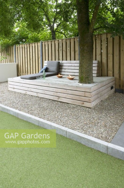 Large wooden lounge bench with cushions and candles under the tree. Path of gravel. Wooden fence.