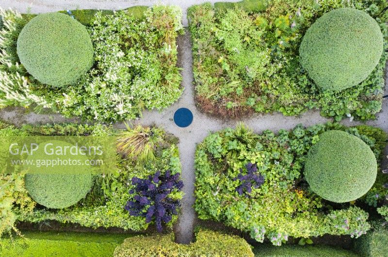 View over rectangular front garden with cruciform gravel paths and birdbath in centre. Mixed border with large mounds of clipped Osmanthus burkwoodii; image taken with drone. September. Summer.