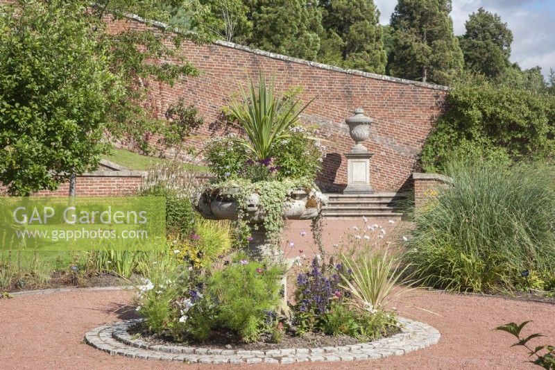 Contemporary, red-brick, walled-garden at Dumfries House. Hard-landscaping. Lawns and plants in flower-beds and borders. Decorative planter on pedestal in circular bed of perennials edged with cobbled stones.  Lidded, stone urn on pedestal. September, Summer.