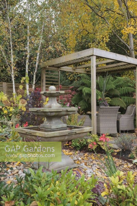 Autumn garden with square tiered stone fountain and wooden pergola under birch trees - October