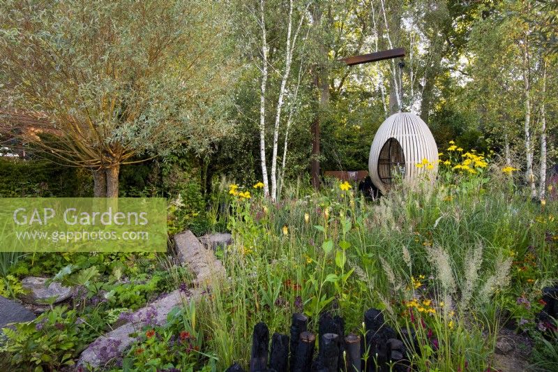 Egg shape swing seat surrounded by Kniphofia 'Tawny King', Rudbeckia laciniata 'Herbstonne', Calamagrostis acutiflora 'Karl Foerster', Calamagrostis brachytricha and Salix Alba. The Yeo Valley Organic Garden. Designer: Tom Massey, supported by Sarah Mead, Chelsea Flower Show 2021. 
