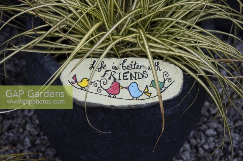 A pot planted with an ornamental grass is brightened up further with a hand painted stone that reads, 'Life is better with friends'. A fun activity to do with children.